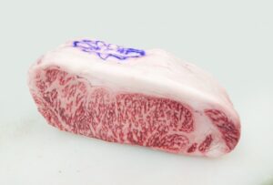 Thick cut of Wagyu beef with delicate marbling and a blue inspection stamp on top, photographed on a light background.